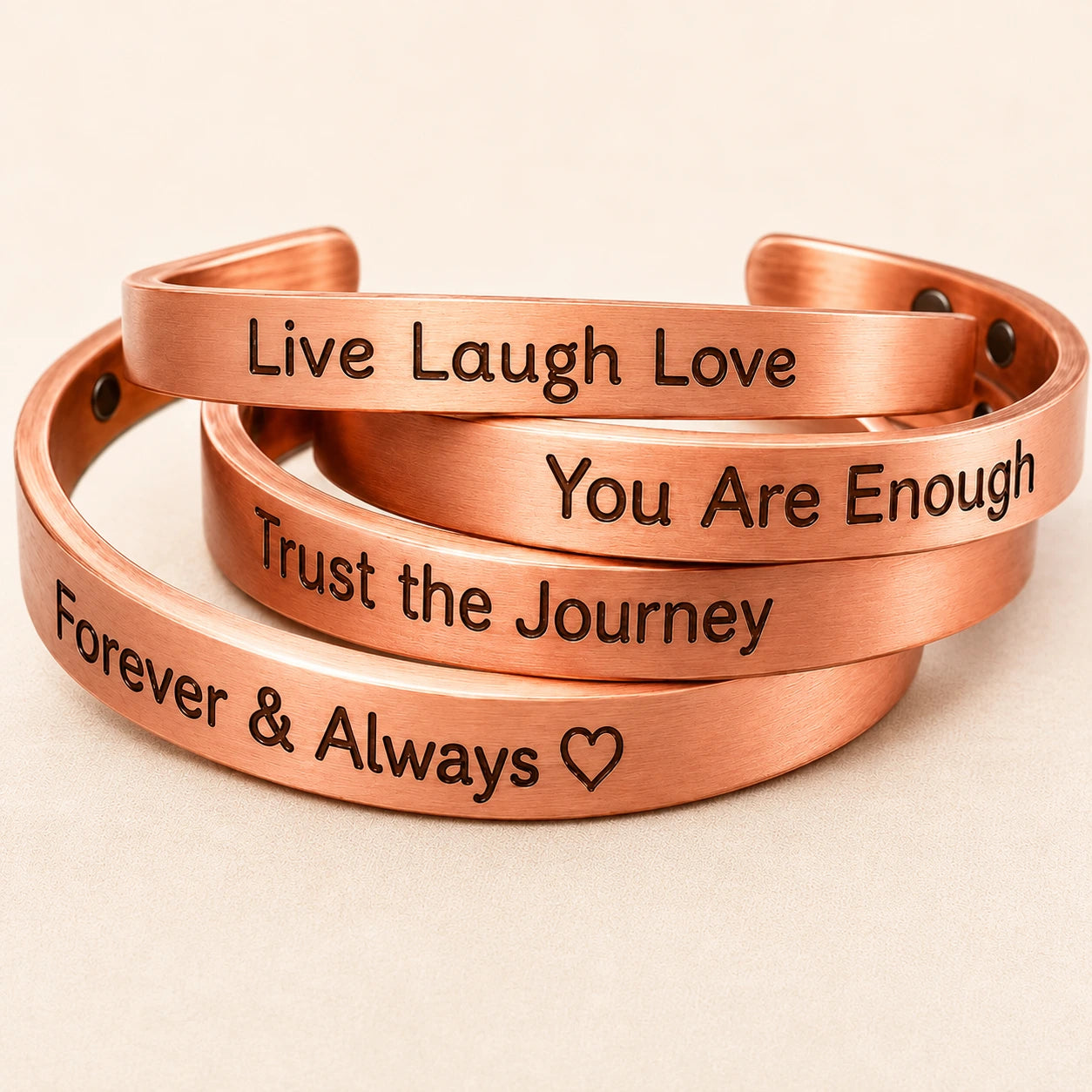Four copper-colored cuff bracelets stacked together on a light beige background, each engraved with a positive affirmation: 'Live Laugh Love,' 'You Are Enough,' 'Trust the Journey,' and 'Forever & Always heart.'.
