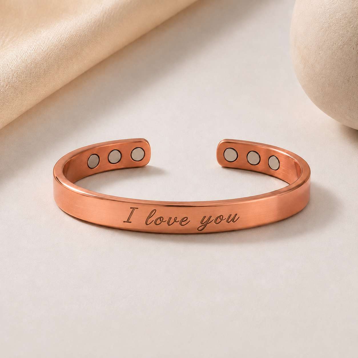 A rose gold cuff bracelet engraved with 'I love you' in script, featuring three circular magnets on each end, displayed on a light beige surface with soft fabric and stone props.