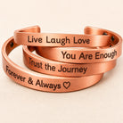 Four copper-colored cuff bracelets stacked together on a light beige background, each engraved with a positive affirmation: 'Live Laugh Love,' 'You Are Enough,' 'Trust the Journey,' and 'Forever & Always heart.'.