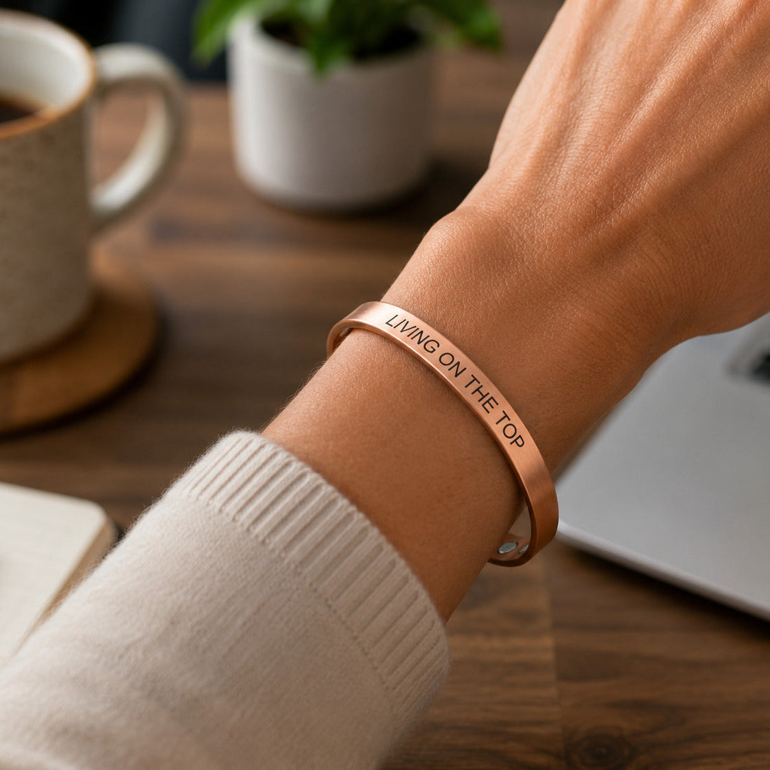 A close-up of a person's wrist wearing a rose gold cuff bracelet engraved with the phrase 'LIVING ON THE TOP'. The background shows a cozy desk setup with a coffee mug, notebook, and laptop.