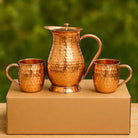 A hammered copper water pitcher with two matching copper mugs displayed on top of a brown cardboard box against a blurred green background.